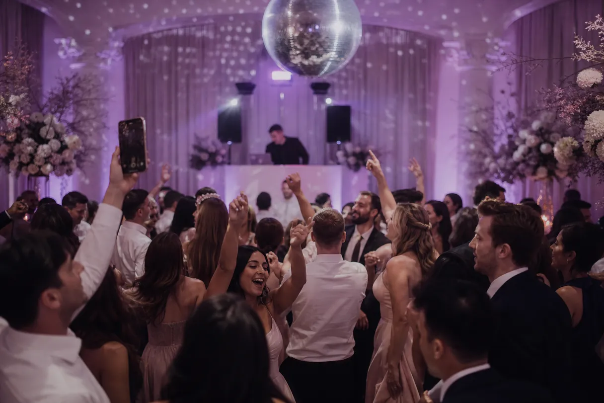 Guests dancing under a disco ball at a wedding reception lit in lilac and plum tones